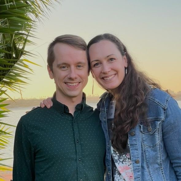 Katie and Lucas Roe smiling together outside at sunset, palm fronds at the edge of the frame
